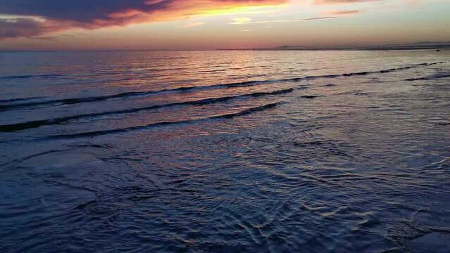 Front view, at ground level of the gentle arrival of the waves on the beach with the sun breaking through clouds in the evening sky