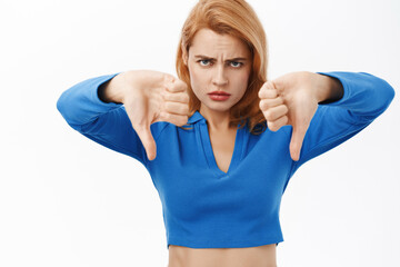 Portrait of angry and upset woman shows thumbs down, do not recommend, dislike smth bad, standing over white background