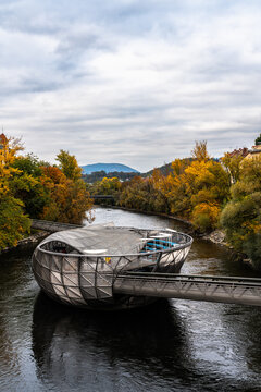 Beautiful view of Murinsel island on the Mur river with colorful trees and mountains in background on a cloudy autumn day, Graz, Austria