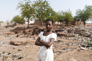 Serious African girl standing on the outskirts of her village surrounded by parched trees, garbage...
