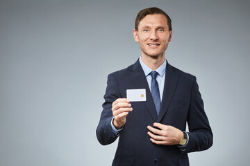 Waist up portrait of smiling businessman showing credit card mockup to camera against grey background, copy space