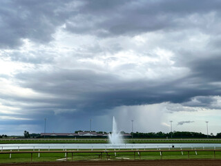 Storm clouds and rain in the background of a fountain in a pond at a horse racetrack.