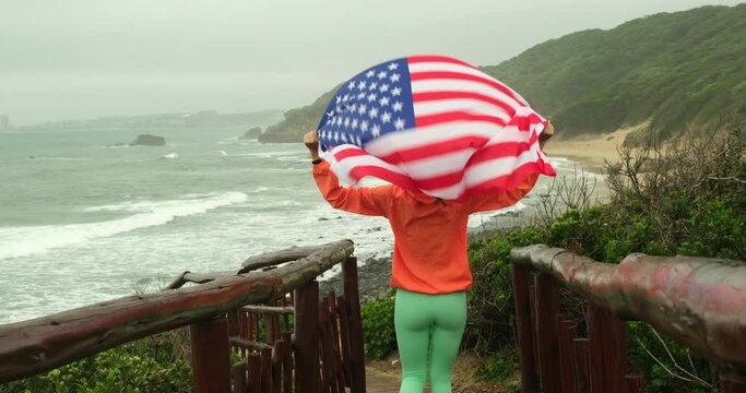 Proud female athlete with USA flag. Athlete in white outfit standing with American flag waving in the wind on the shore. athletic girl runs on a wooden bridge near the ocean in autumn