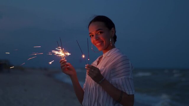 African american woman smiling and posing on night party at seaside with sparklers lights in hands on Fourth of July. Pretty girl portrait partying on beach with fireworks on New Year, Christmas.