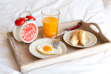 A red alarm clock, a glass of orange juice and toast are on a wooden tray that stands on a white bed.