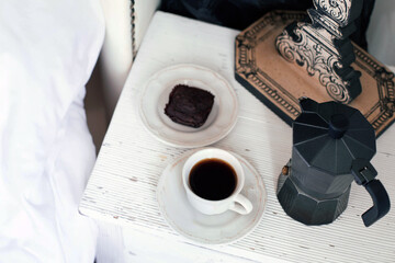 A white cup of black coffee, a coffee pot and a chocolate cake are on the bedside table near the white bed. The theme of breakfast with coffee. 