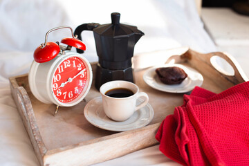 Theme breakfast with coffee. A tray with a red alarm clock, a coffee pot and a cup of black coffee stands on a white bed illuminated by sunlight.