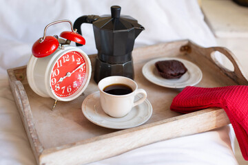 Theme breakfast with coffee. A tray with a red alarm clock, a coffee pot and a cup of black coffee are on a white bed in the morning.