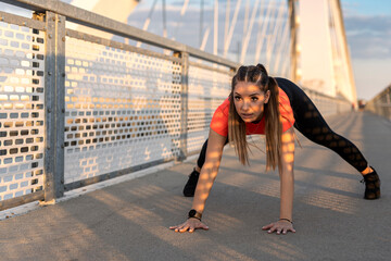 Fototapeta premium Young happy focused fitness girl in black yoga pants and orange short shirt work out and stretch her body on the bridge footpath during the day. Front view.