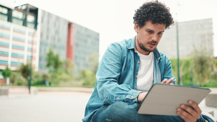 young bearded man in denim shirt sitting and using tablet outdoors on modern buildings background