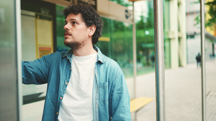 Close-up, young bearded man in denim shirt stands at bus stop and looks at the timetable on modern...