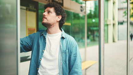 Close-up, young bearded man in denim shirt stands at bus stop and looks at the timetable on modern city background