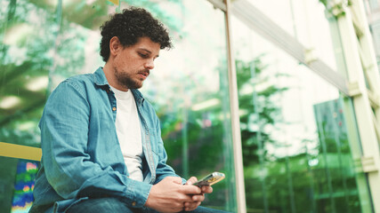 Close-up, young bearded man in denim shirt sits at bus stop waiting for bus and uses mobile phone...