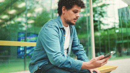Close-up, young bearded man in denim shirt sits at bus stop waiting for bus and uses mobile phone on modern city background