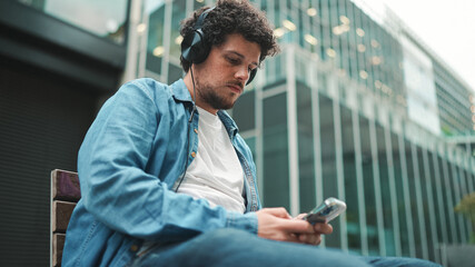 Close-up of young bearded man in denim shirt sitting in headphones on bench listening to music and using mobile phone on modern buildings background