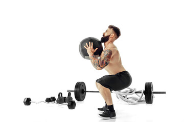 Muscular tattooed bearded male exercising fitness weights Medicine Ball in studio isolated on white...