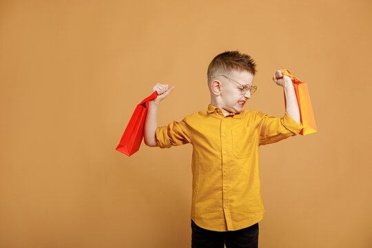 Image of smiling boy holding bags with presents or shoppings on yellow background. child in glasses lifts packages like weights