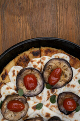 Vegan pizza. Top view of a vegetarian pizza with farinata, vegan mozzarella cheese, tomatoes, grilled eggplant and fresh oregano in a black pizza pan on the wooden table.