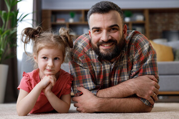 Family portrait at home. Young handsome father with cute daughter lying down on a carpet near the couch, looking at camera and smiling. Father's day