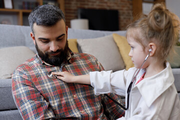 Cute small girl playing doctor with her father. Funny child holds stethoscope, listening to father patient at home. Happy family enjoy spending time together.