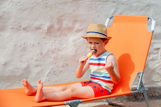 Little Boy Sit In Orange Sunbed And Eats Ice-cream