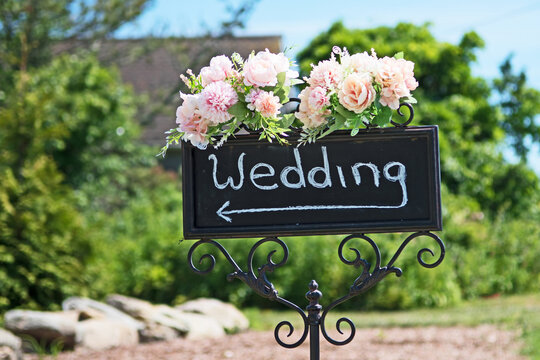 A directional sign for a wedding topped by beautiful pink flowers and with a blurred background.