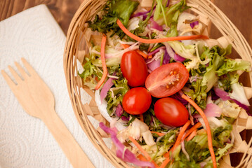 Red cherry tomatoes in a wicker plate with salad. Vegetarian dish of fresh lettuce, arugula, carrots, red cabbage and tomatoes.
