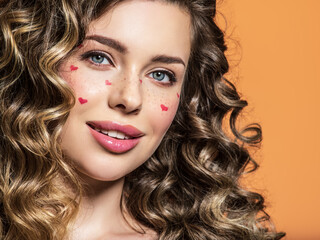 Portrait of a beautiful smiling girl with painted red hearts on her face. Fashion photography. Sexy blonde with curly hair. An expressive and sensual model with bright makeup poses in the studio.