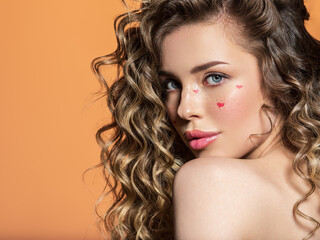 Portrait of a beautiful girl with painted red hearts on her face. Fashion photography. Sexy blonde with curly hair. An expressive and sensual model with bright makeup poses in the studio.