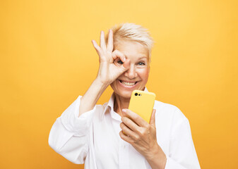 Happy mature senior woman with short hair holding smartphone using mobile and show finger ring...
