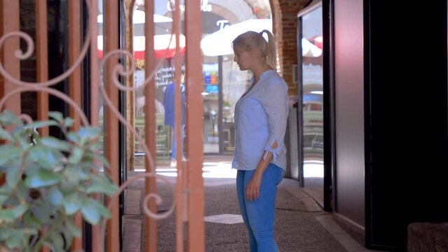 Behind a red barrier, a blonde Caucasian woman walks up, stops to take out her smartphone and take a picture of the window on the left