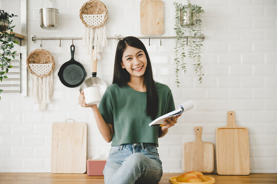 Happy young asian woman sitting on counter kitchen at home. Female reading note cooking