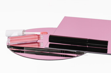 Lip gloss, mascara, and an eye brow brush on a mirror with a pink reflection. 