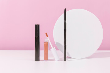 Mascara, lip gloss and a backup brush in front of a pink background and a circle shape.