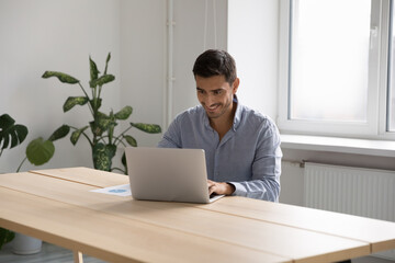 Man office employee work on laptop sit at desk in cozy workplace use corporate program looks satisfied by work results, e-mailing to client, search helpful information on internet. Modern tech concept