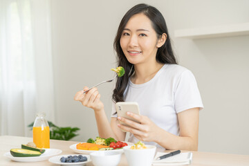 Dieting, asian young woman eating, holding fork at broccoli, diet plan nutrition with fresh vegetables salad, enjoy meal while using smartphone. Nutritionist of healthy, nutrition of weight loss.