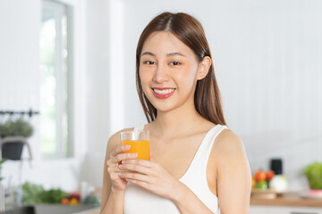 Happy housewife, smile asian young woman, girl drinking fresh orange juice in a glass drink in the kitchen, holding transparent glass in her hand, thirsty at home. Health care, healthy diet concept.