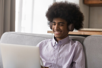 Cheerful teenage 18s African guy wear headphones sit on sofa with laptop, spend leisure online,...