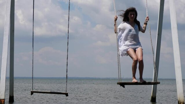 A young woman in a white sundress swings on a swing against the background of the sea. Swing over water. 4K slow motion.