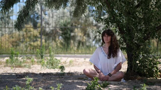 A young woman meditates under a tree in a white sundress.