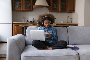 Smiling African student girl studying alone at home use laptop, listen audio task through wireless...
