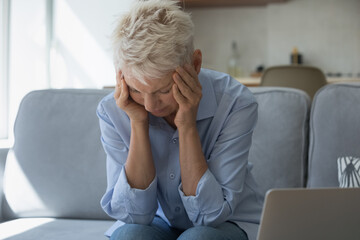 Upset stressed older woman holds head with hands sit on sofa near laptop, suffers from strong severe headache, chronic migraine after device usage, information overload, awful news, bank debt concept
