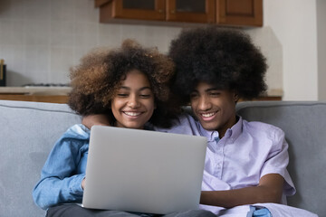 Obraz na płótnie Canvas Cheerful African girlfriend and boyfriend, teenager couple sit on sofa with laptop, spend leisure at home watch on-line movie on digital streaming TV services, young gen modern tech usage, fun concept