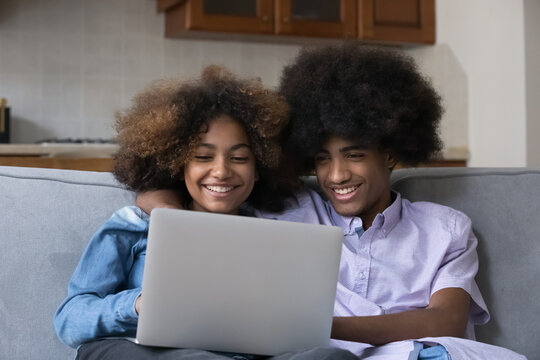 Cheerful African girlfriend and boyfriend, teenager couple sit on sofa with laptop, spend leisure at home watch on-line movie on digital streaming TV services, young gen modern tech usage, fun concept