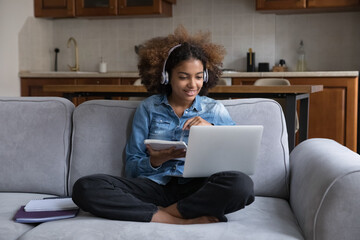 Cute teenage girl wear headphones makes assignment sit on sofa with laptop, gaining new knowledge...