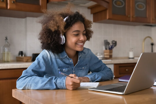 Pretty African girl wear headphones studying sit at table in kitchen use laptop engaged in distancing class with on-line tutor smile enjoy learning feels motivated. Education, video call event concept
