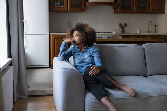 Sad African teen girl sit on sofa lost in thoughts, thinks staring into distance, looks upset experiences first unrequited love, having low self-esteem, problems, feels insecure. Teen problem concept
