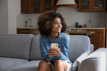 Cute African teenage girl resting on cozy sofa holds smartphone staring aside looks dreamy,...