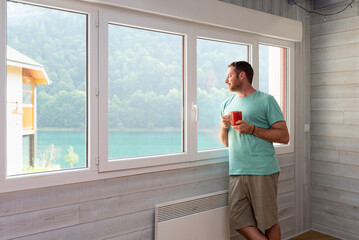 Relaxed man holding a mug and enjoying a lake views from the window of his apartment.