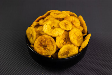Fried banana chips or banana wafers, arranged beautifully in a black ceramic bowl with a black textured background.